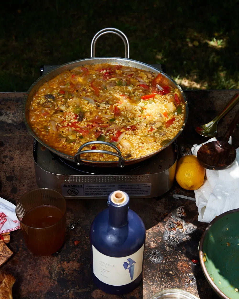 A large pan of paella filled with rice, seafood, and vegetables sits on an outdoor stove. Surrounding the pan are a blue bottle, a glass of amber liquid, a lemon, a napkin, and a green plate on a rustic tabletop.