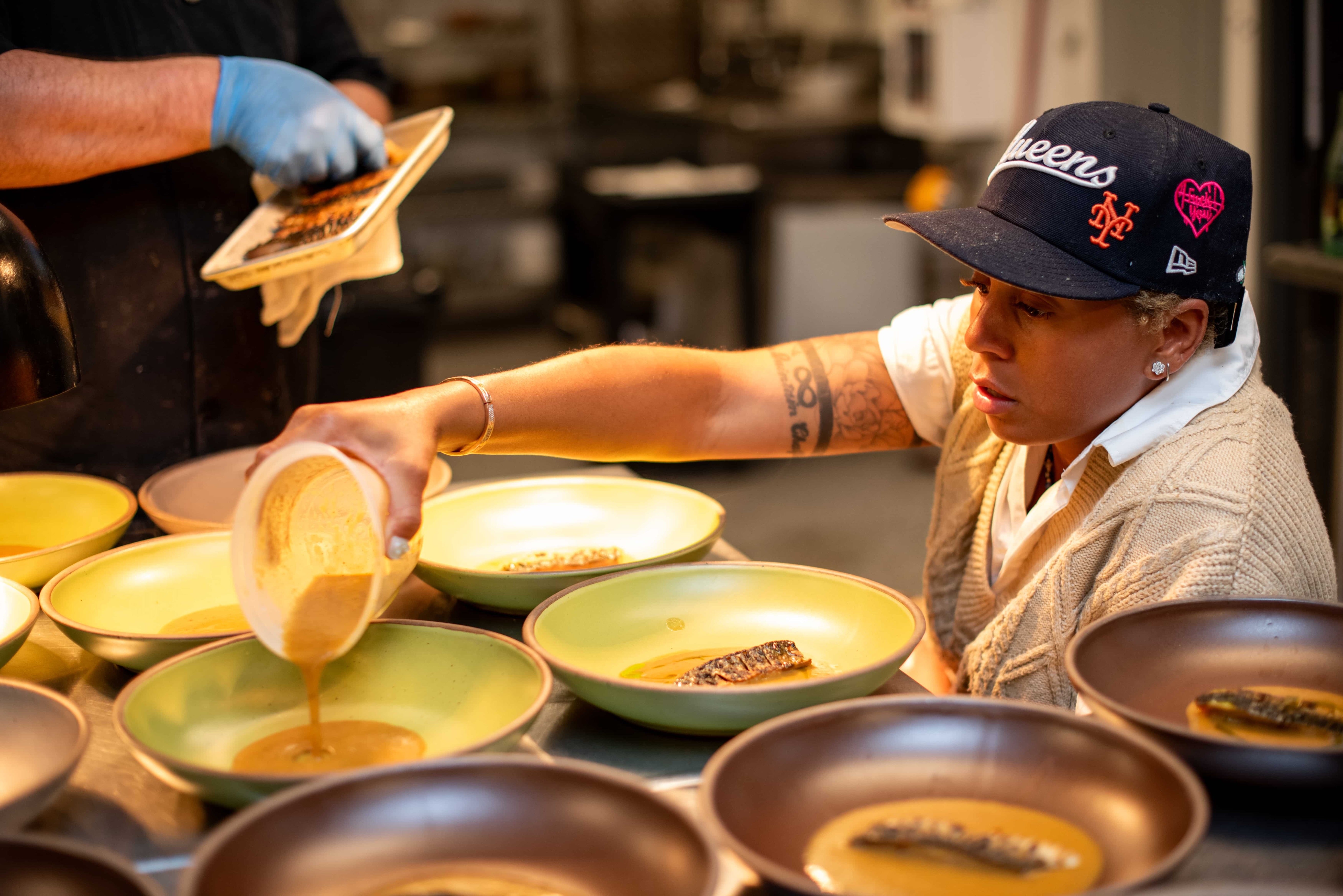 Lay Alston pouring a sauce as the base for a dish into various ceramic bowls while someone else adds to the dish.