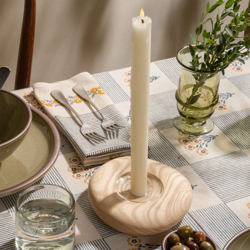 Lit white taper candle in a light wooden holder on a patterned table setting with glassware and olives.