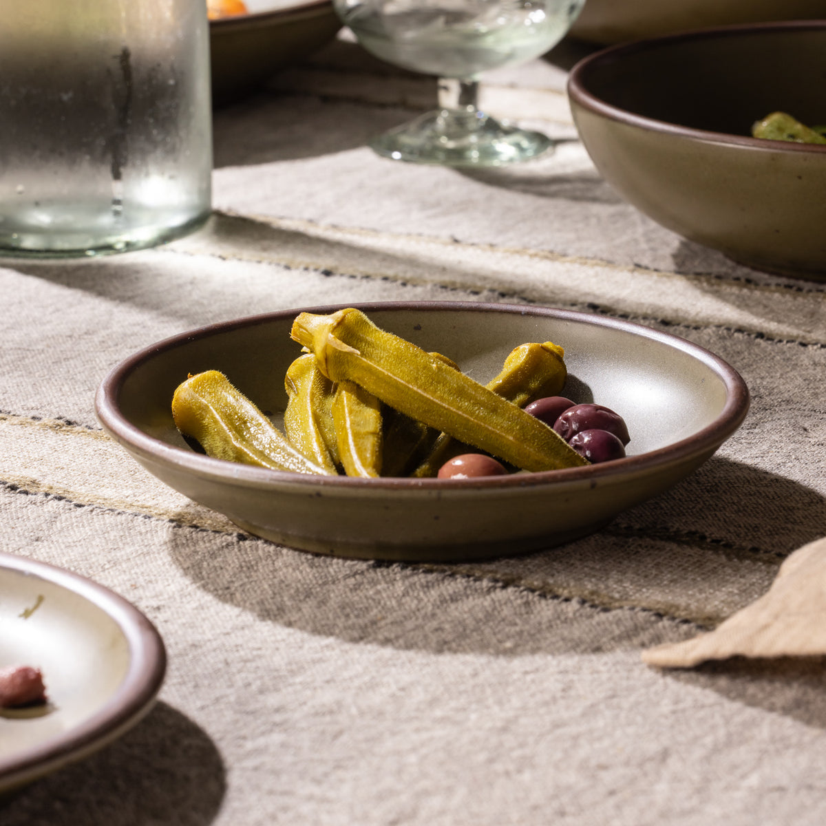 Shallow olive-toned stoneware dish filled with pickled okra and olives, styled on a rustic linen-covered table.