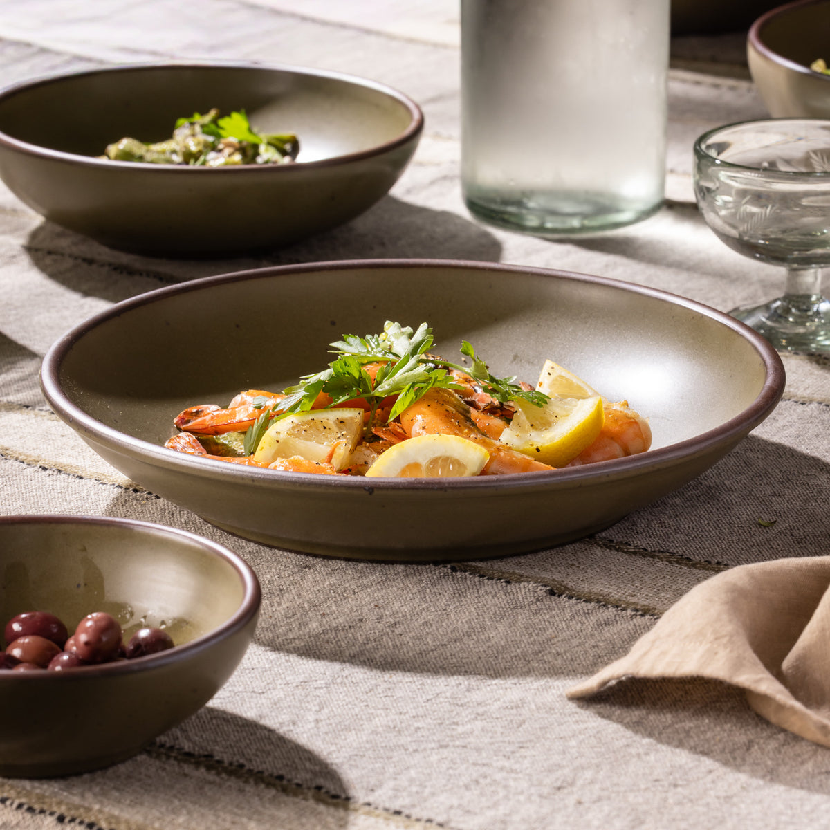 Olive-toned stoneware coupe plate with shrimp and lemon, shown alongside coordinating tableware.