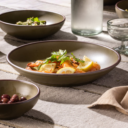 Olive-toned stoneware coupe plate with shrimp and lemon, shown alongside coordinating tableware.