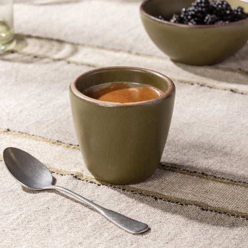 Olive-toned stoneware cup filled with a hot beverage, shown with a spoon on a striped linen tablecloth.