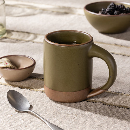 Olive-glazed stoneware mug with exposed clay base and sculpted handle, styled with tea on a rustic linen table.