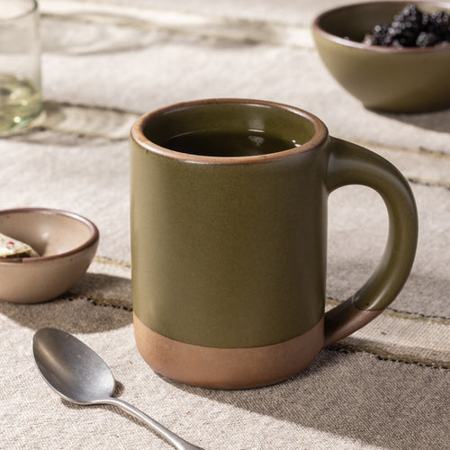 Tall olive-glazed stoneware mug with exposed clay base and curved handle, styled with coffee on a rustic linen table.