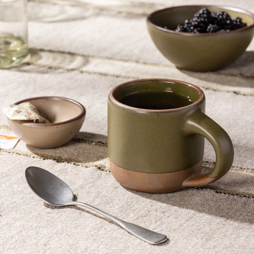 Short olive-toned stoneware mug with a raw clay base, shown with tea and coordinating tabletop pieces.