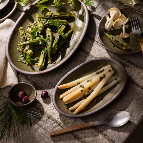 Olive-toned stoneware oval platters and plates, styled with seasonal vegetables and shared dishes on a rustic linen table.