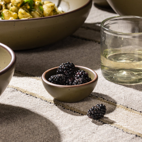Small stoneware bowl filled with blackberries, shown on a striped linen tablecloth beside a drinking glass.