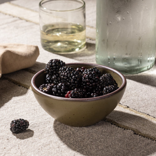 Small stoneware bowl filled with blackberries, shown on a striped linen tablecloth beside a glass and carafe.