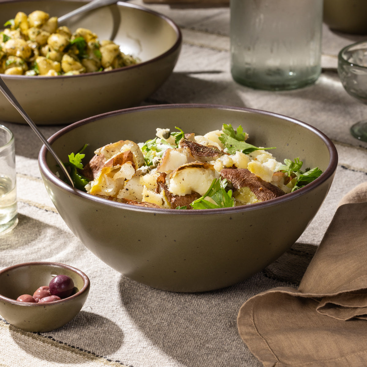 Large stoneware serving bowl filled with potato salad, shown on a striped linen tablecloth with a serving spoon.