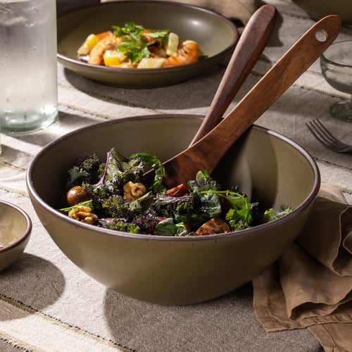 Large stoneware serving bowl filled with mixed greens and tomatoes, shown with wooden salad servers on a striped linen tablecloth.