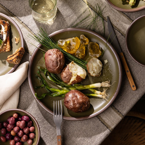 Olive-toned stoneware dinner plate, styled with roasted potatoes, grilled scallions, and seasonal accompaniments on a rustic linen table.