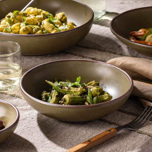 Olive-toned stoneware shallow dinner bowl with grilled okra, shown alongside coordinating dinnerware and glassware.