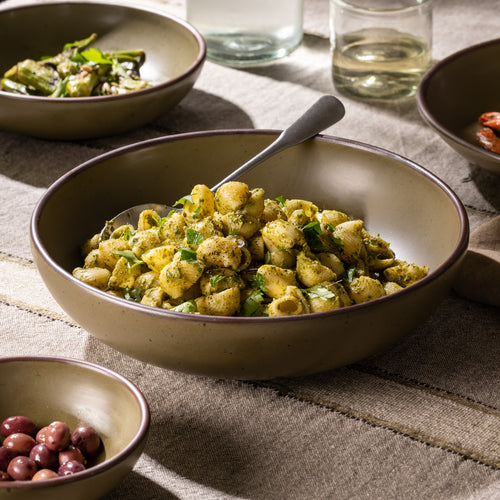 Large stoneware serving bowl filled with shell pasta in green herb sauce, shown on a striped linen tablecloth.