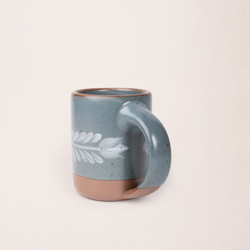 Side profile of Blue-grey ceramic mug with a hand-painted white leaf motif and exposed clay base on a plain white background.