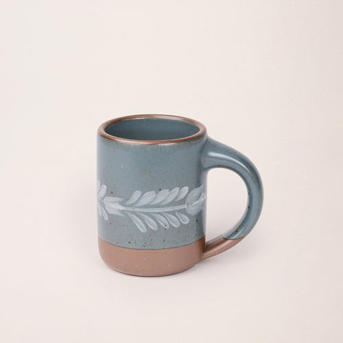 Blue-grey ceramic mug with a hand-painted white leaf motif and exposed clay base on a plain white background.