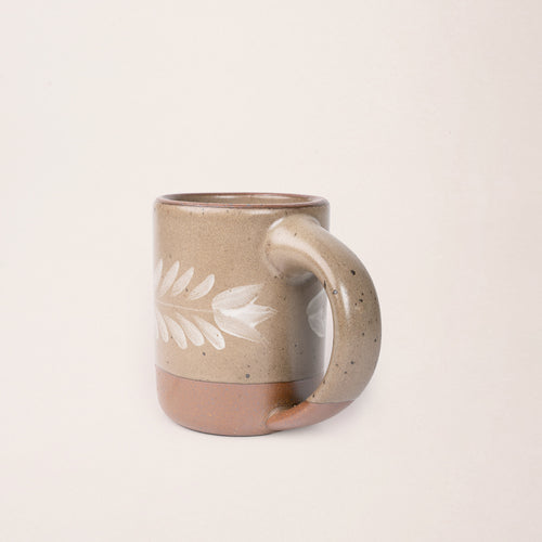 Side profile of a pale warm brown ceramic mug with a hand-painted white leaf motif and exposed clay base on a plain white background.