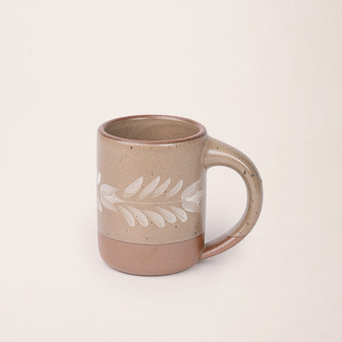 Pale warm brown ceramic mug with a hand-painted white leaf motif and exposed clay base on a plain white background.