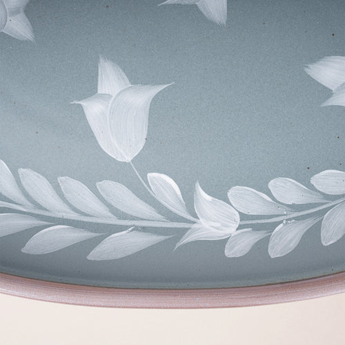 Closeup of an Oval ceramic platter with a hand-painted white leaf and flower border on a blue-grey background with an exposed clay rim.