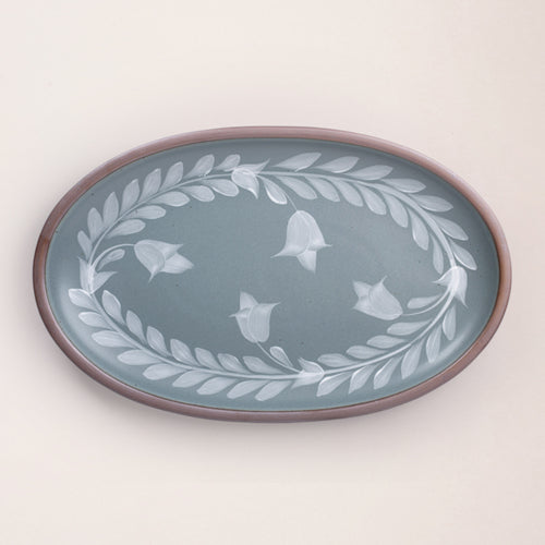 Oval ceramic platter with a hand-painted white leaf and flower border on a blue-grey background with an exposed clay rim.
