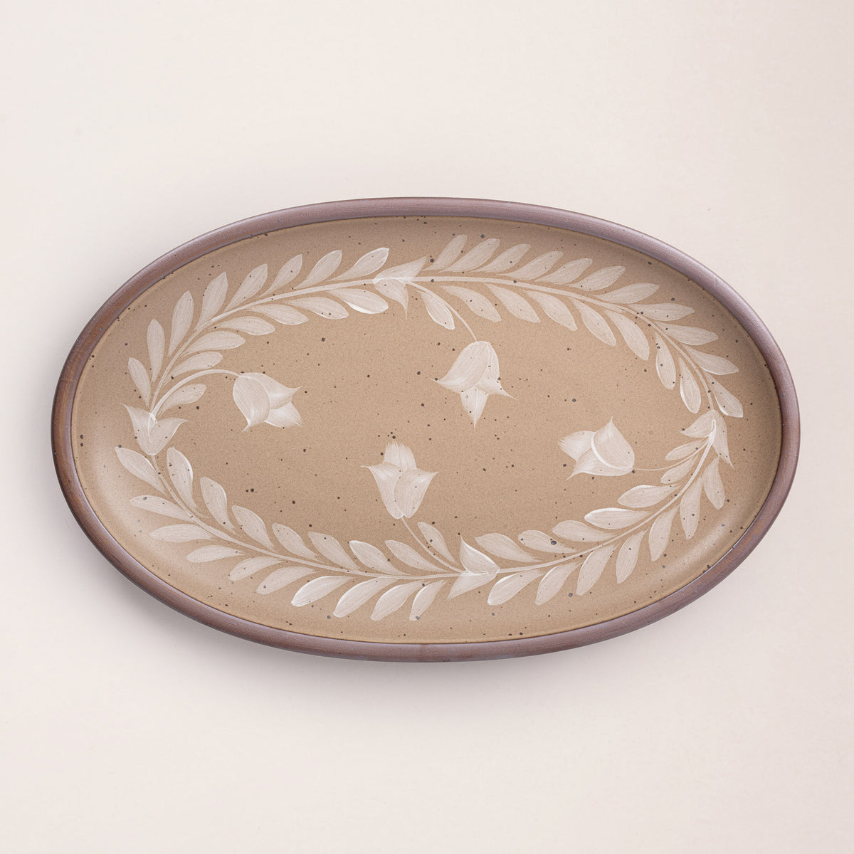 Oval ceramic platter with a hand-painted white leaf and flower border on a warm pale brown background with an exposed clay rim.