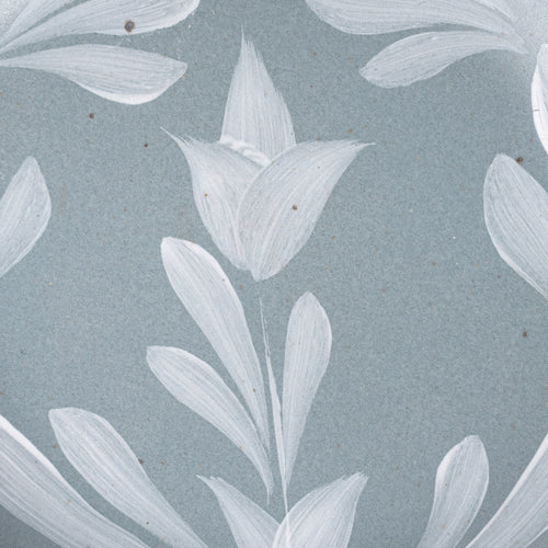 Closeup of a small round ceramic plate with a hand-painted white flower and leaf design on a blue-grey background with an exposed clay rim.