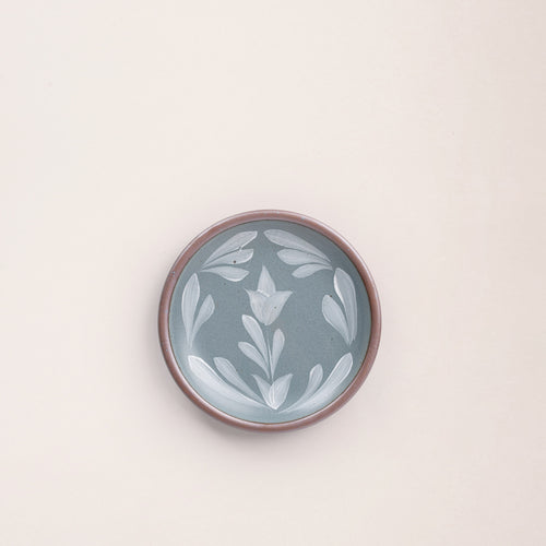 Small round ceramic plate with a hand-painted white flower and leaf design on a blue-grey background with an exposed clay rim.