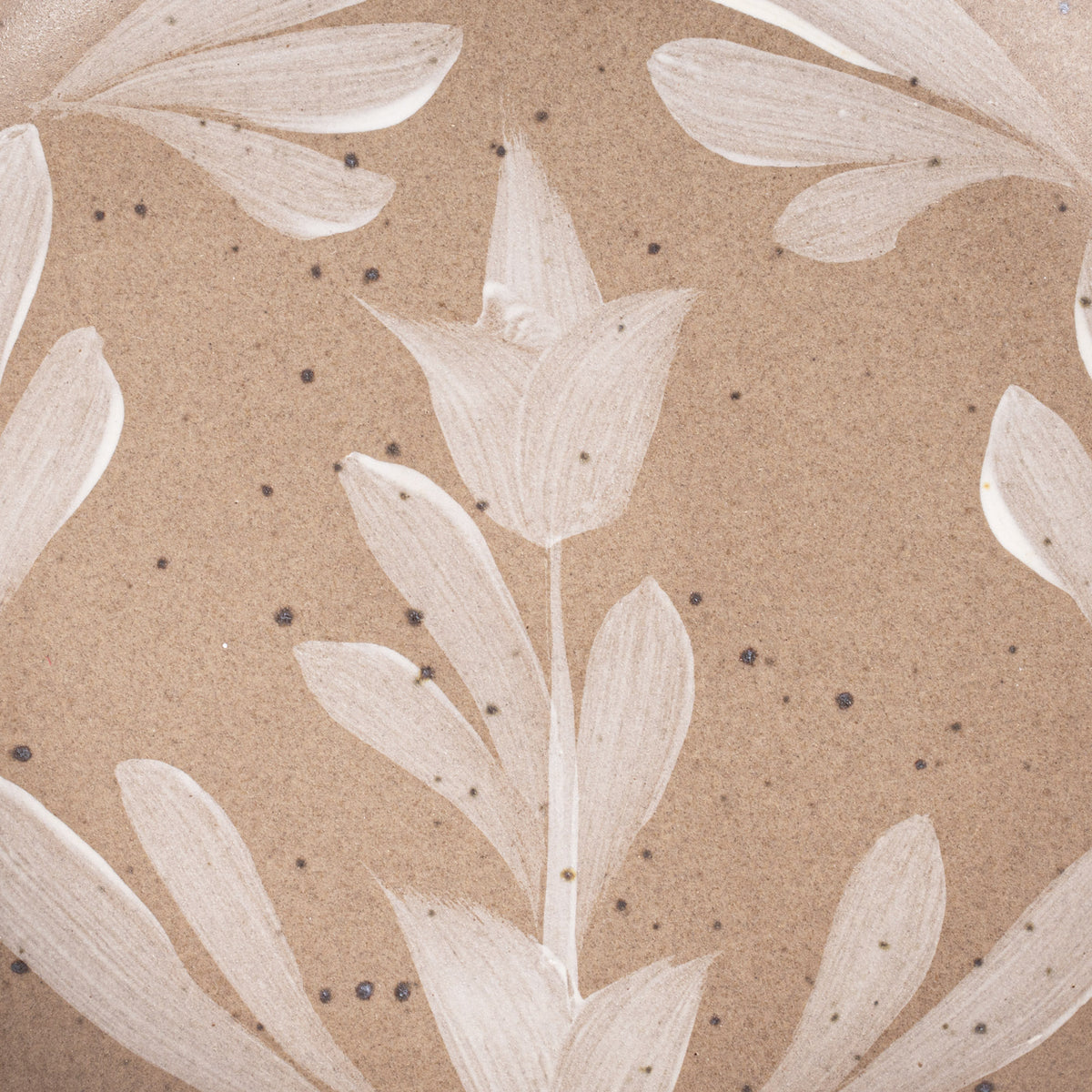 Closeup of Small round ceramic plate with a hand-painted white flower and leaf design on a warm pale brown background with an exposed clay rim.