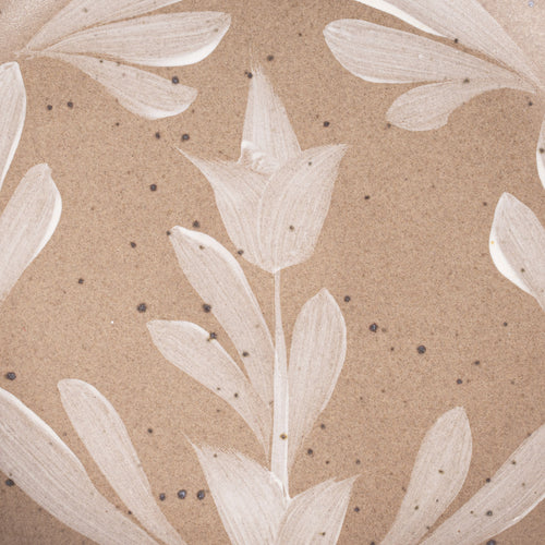 Closeup of Small round ceramic plate with a hand-painted white flower and leaf design on a warm pale brown background with an exposed clay rim.