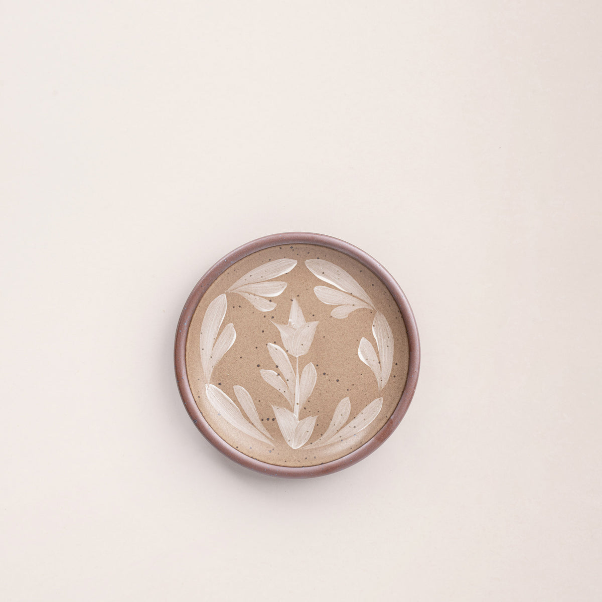 Small round ceramic plate with a hand-painted white flower and leaf design on a warm pale brown background with an exposed clay rim.