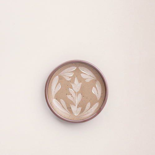 Small round ceramic plate with a hand-painted white flower and leaf design on a warm pale brown background with an exposed clay rim.