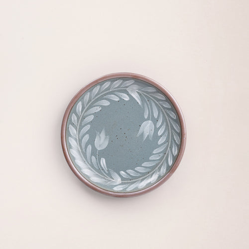 Side round ceramic plate with a hand-painted white leaf wreath and floral design on a blue-grey background with an exposed clay rim.
