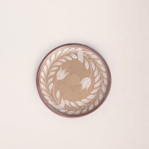Side round ceramic plate with a hand-painted white leaf wreath and floral design on a warm pale brown background with an exposed clay rim.