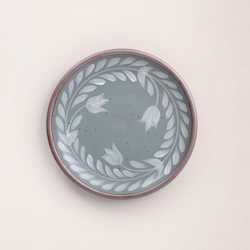 Dinner round ceramic plate with a hand-painted white leaf wreath and floral design on a blue-grey background with an exposed clay rim.