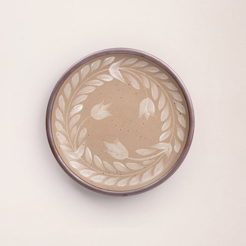Dinner round ceramic plate with a hand-painted white leaf wreath and floral design on a warm pale brown background with an exposed clay rim.