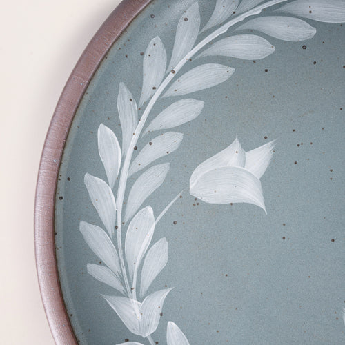 Closeup of a large round ceramic platter with a hand-painted white leaf wreath and floral design on a blue-grey background with an exposed clay rim.