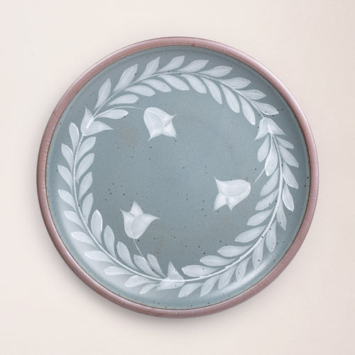 Large round ceramic platter with a hand-painted white leaf wreath and floral design on a blue-grey background with an exposed clay rim.