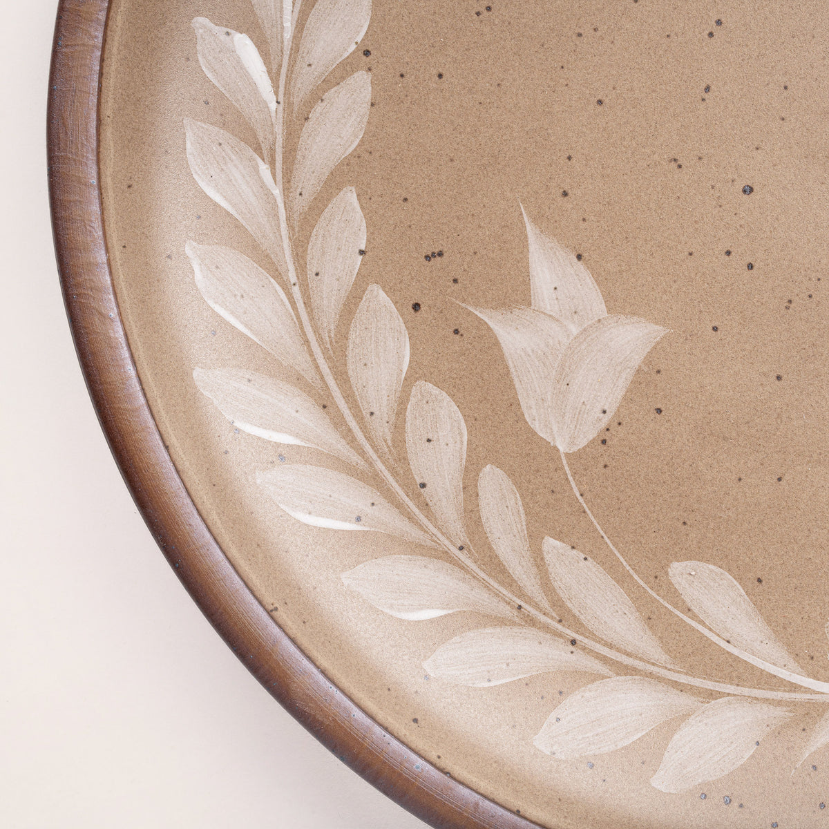 Closeup of a large round ceramic platter with a hand-painted white leaf wreath and floral design on a warm pale brown background with an exposed clay rim.