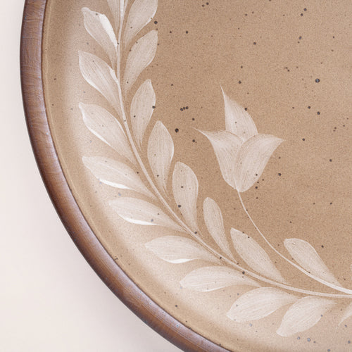 Closeup of a large round ceramic platter with a hand-painted white leaf wreath and floral design on a warm pale brown background with an exposed clay rim.