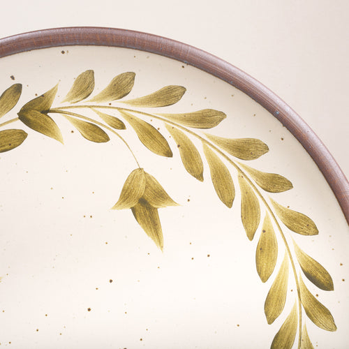 Closeup of a large round ceramic platter with a hand-painted olive green leaf wreath and floral design on a cream background with an exposed clay rim.