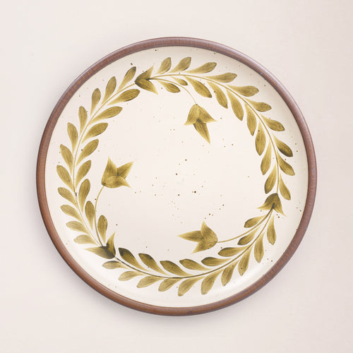 Large round ceramic platter with a hand-painted olive green leaf wreath and floral design on a cream background with an exposed clay rim.
