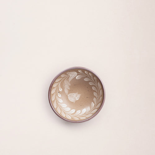 Overhead view of a small ceramic shallow bowl with a hand-painted white leaf border on a pale warm brown interior and exterior with an exposed clay rim.