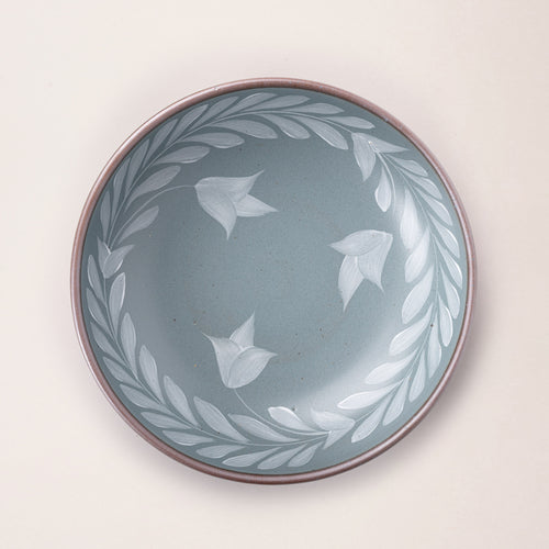 Overhead view of Large ceramic shallow bowl with a hand-painted white leaf border on a blue-grey interior and exterior with an exposed clay rim.