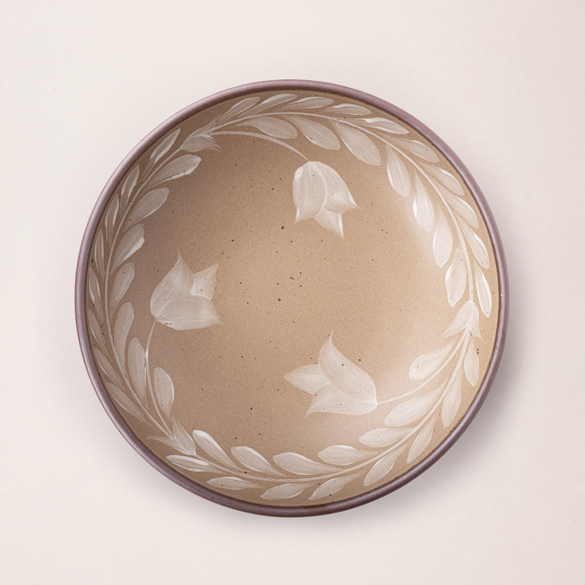 Overhead view of a large ceramic shallow bowl with a hand-painted white leaf border on a pale warm brown interior and exterior with an exposed clay rim.