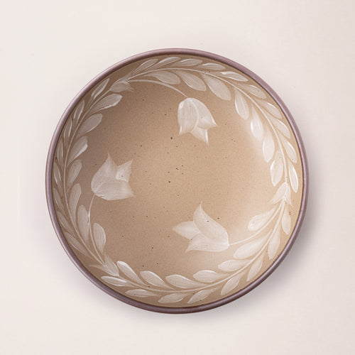 Overhead view of a large ceramic shallow bowl with a hand-painted white leaf border on a pale warm brown interior and exterior with an exposed clay rim.