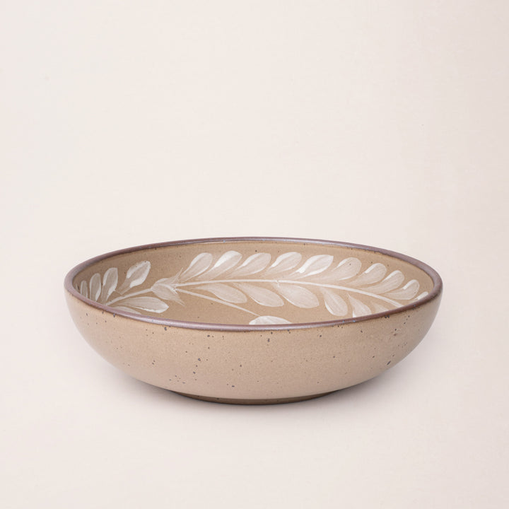 Large ceramic shallow bowl with a hand-painted white leaf border on a pale warm brown interior and exterior with an exposed clay rim.