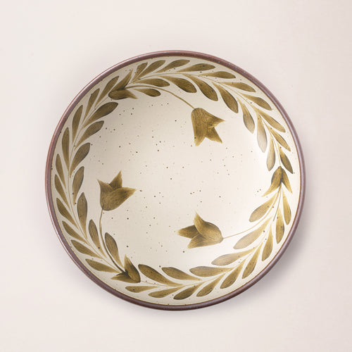 An overhead view of a large ceramic shallow bowl with a hand-painted olive green leaf border on a cream interior and exterior with an exposed clay rim.