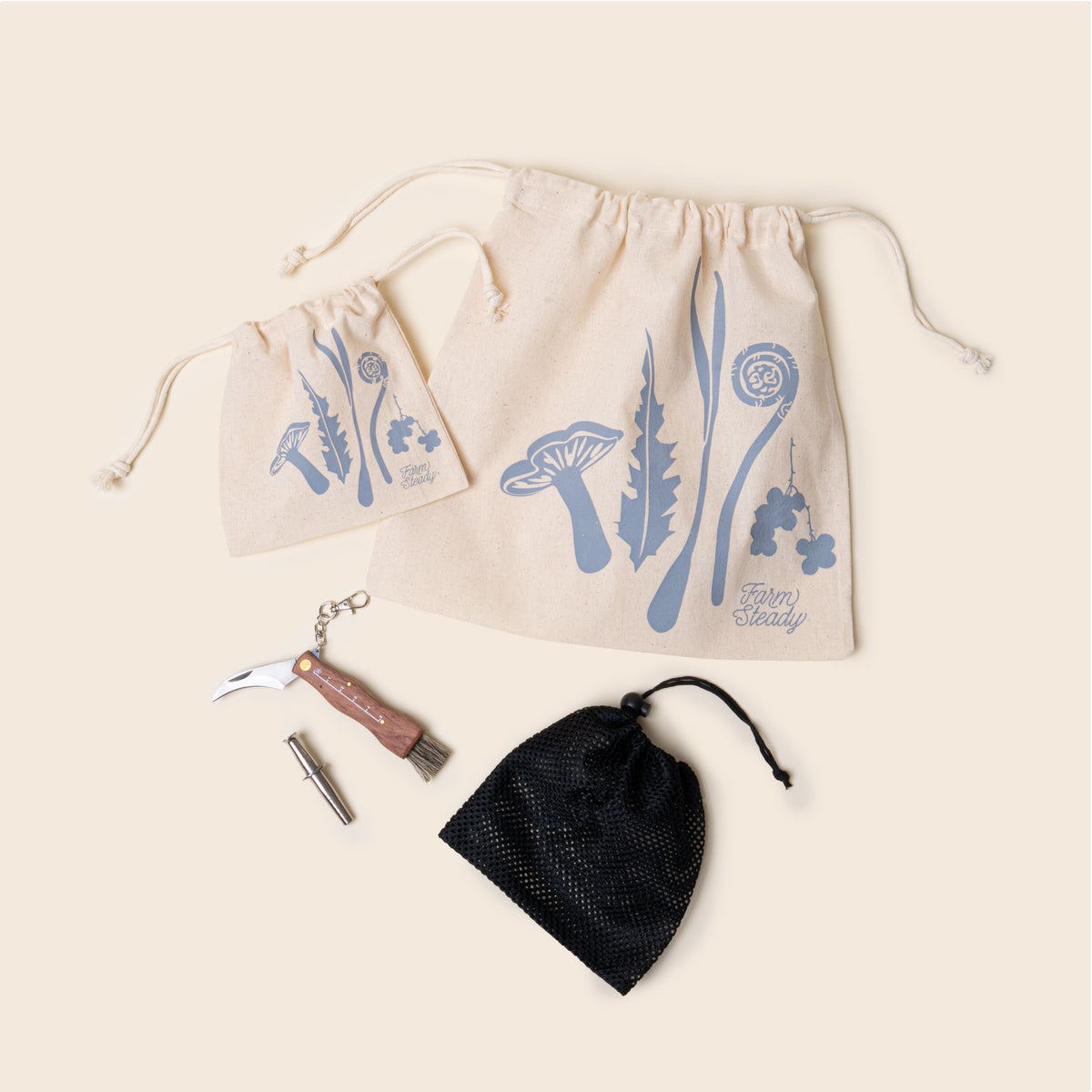 Flat lay of the FarmSteady Foraging Tool Kit contents — two cream drawstring bags with blue plant illustrations, a black mesh bag, a folding foraging knife with a wooden handle and brush, and a small silver tool.