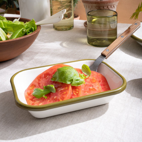 Tomato slices and basil with a fork in a small rectangular baking dish in a white color with an olive green rim.