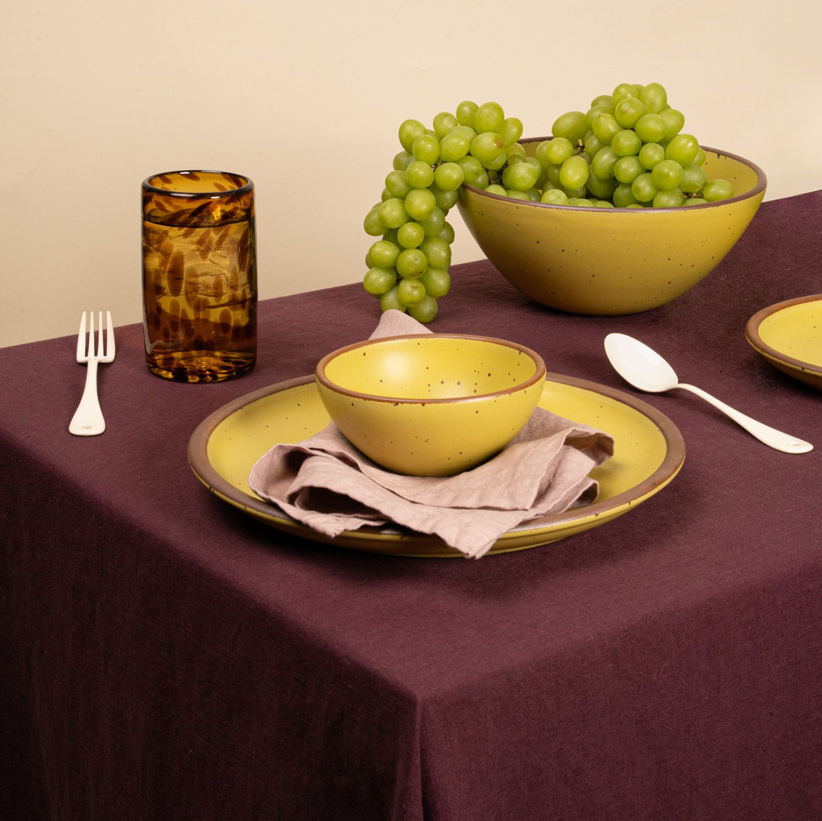 A dinner setting featuring speckled yellow ceramic bowls and plates on a deep purple tablecloth, with a sprig of green grapes in a larger bowl and an amber glass next to silverware. A beige napkin is placed under the smaller bowl.